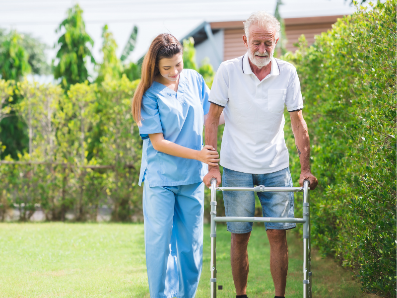 A young female caregiver in blue scrubs helps an elderly man using a walker as they walk together outside on a sunny day, surrounded by green bushes and trees.