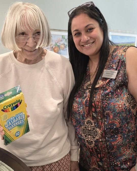 Two women smiling together indoors; one holds a pack of Crayola markers, while the other wears a name tag. Artworks are visible on the wall behind them.