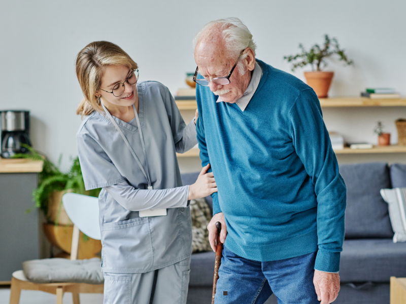 A young caregiver in scrubs assists an elderly man with glasses and a cane as he walks in a living room, offering support with a gentle smile.