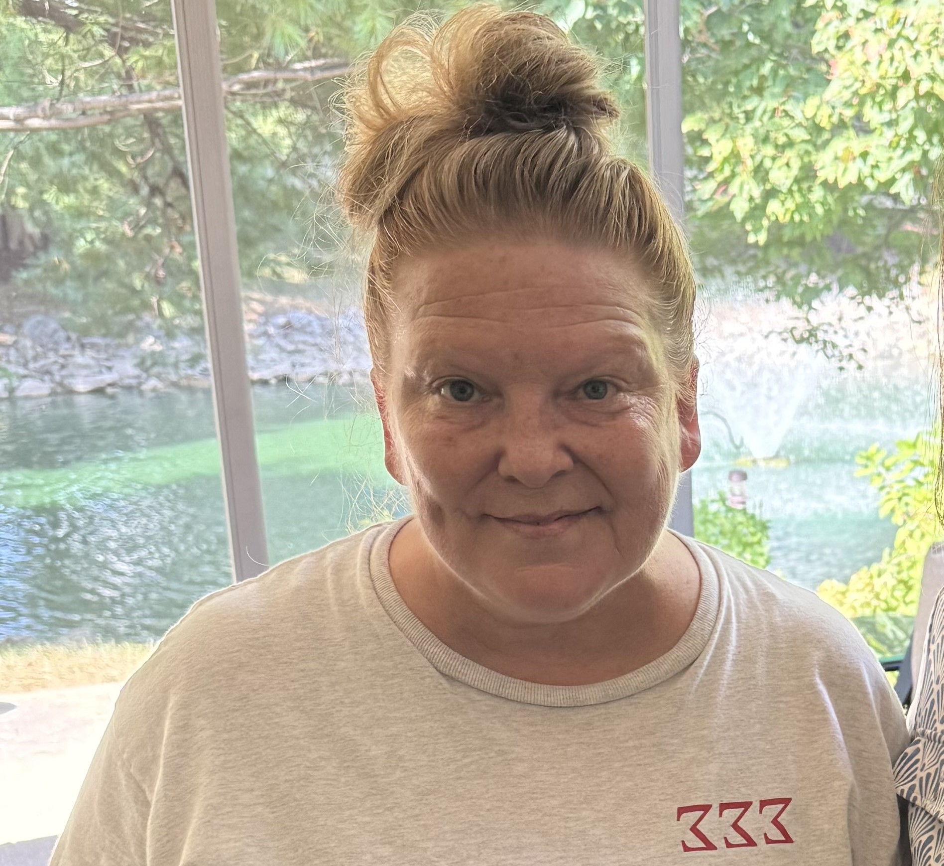 A woman with blonde hair in a bun, wearing a light-colored shirt with 333 in red, stands indoors by a window with a leafy green outdoor scene and water visible behind her.