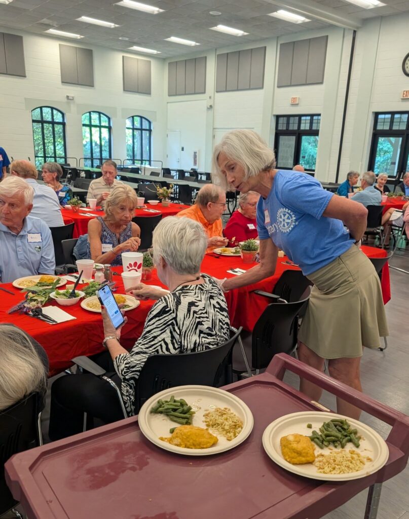 A woman in a blue shirt and khaki skirt speaks to a seated woman at a community meal event with red tablecloths. In the foreground, two plates of food rest on a brown tray.
