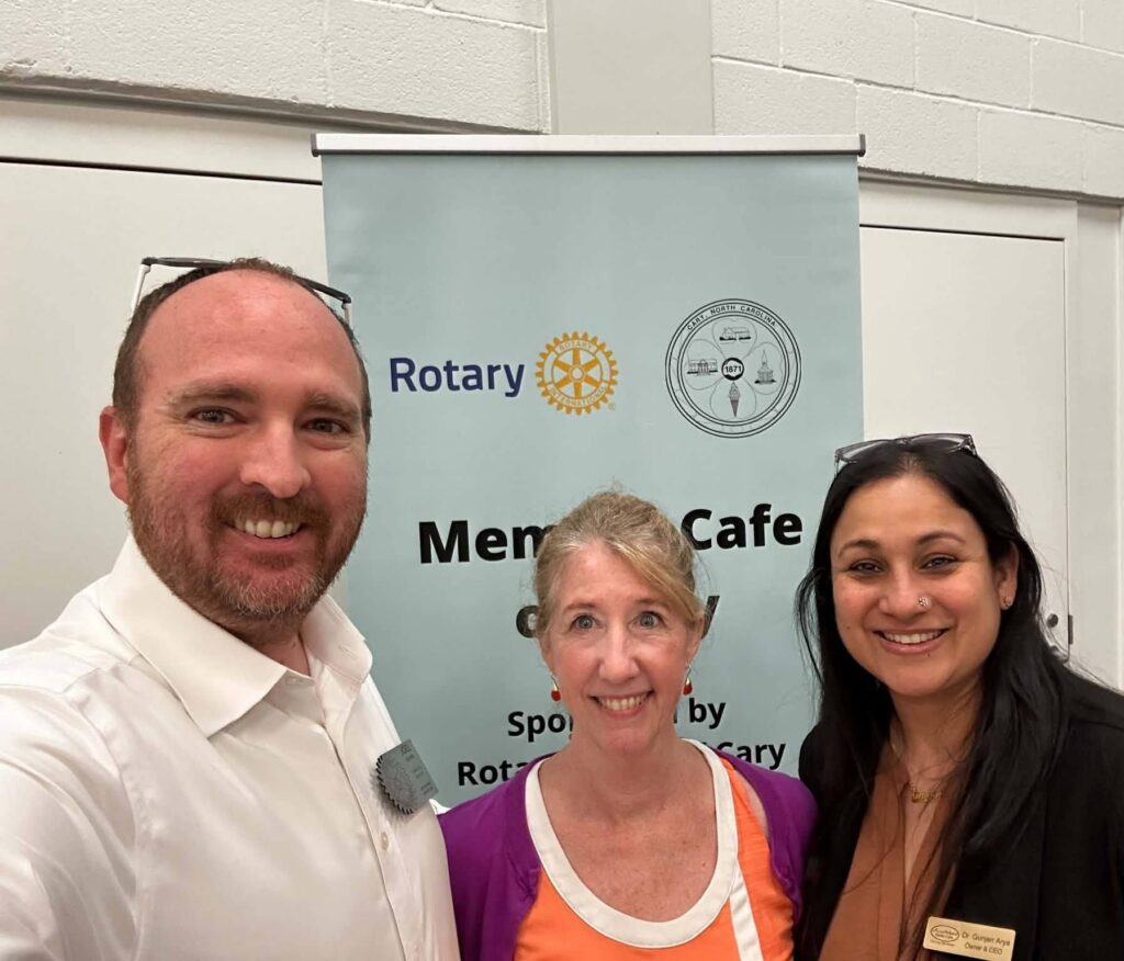 Three people smiling in front of a Memory Cafe banner sponsored by Rotary clubs. The group is indoors, wearing business-casual attire, and posing for a photo at an event.