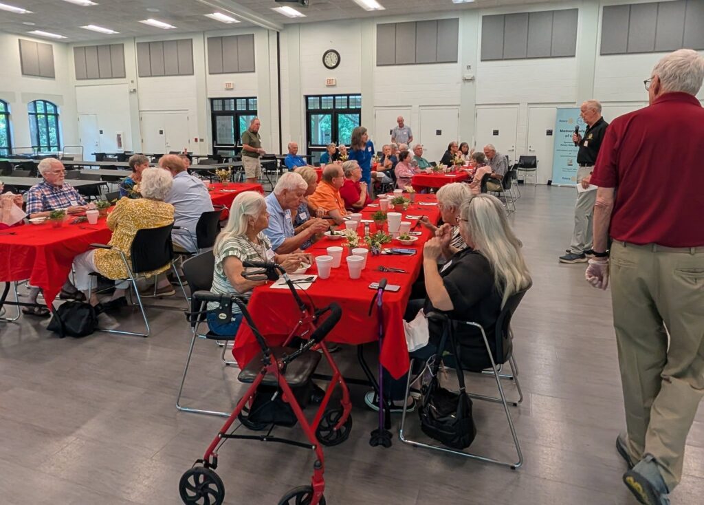A group of elderly people gathers around tables with red tablecloths in a community hall, eating and socializing. A red walker is beside one table. The room has bright lighting and large windows.