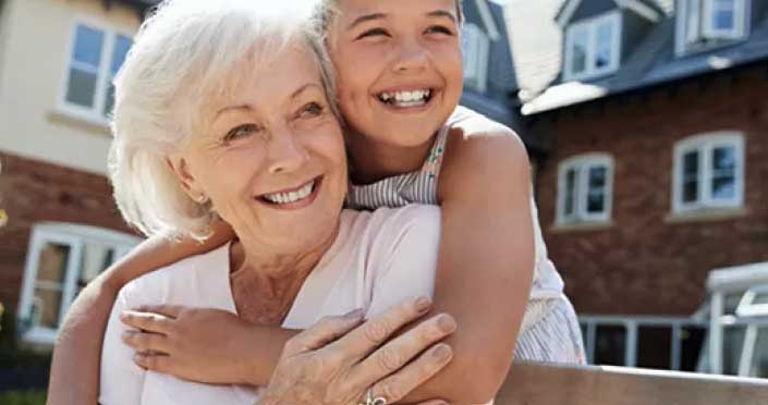 Smiling older woman with white hair sits on a bench while a happy young girl hugs her from behind. Both are outdoors in front of a brick house.