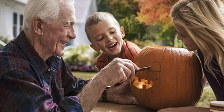 An elderly man carves a pumpkin with two smiling children, a boy and a girl, outdoors on a sunny day. The pumpkin has a partially carved face.
