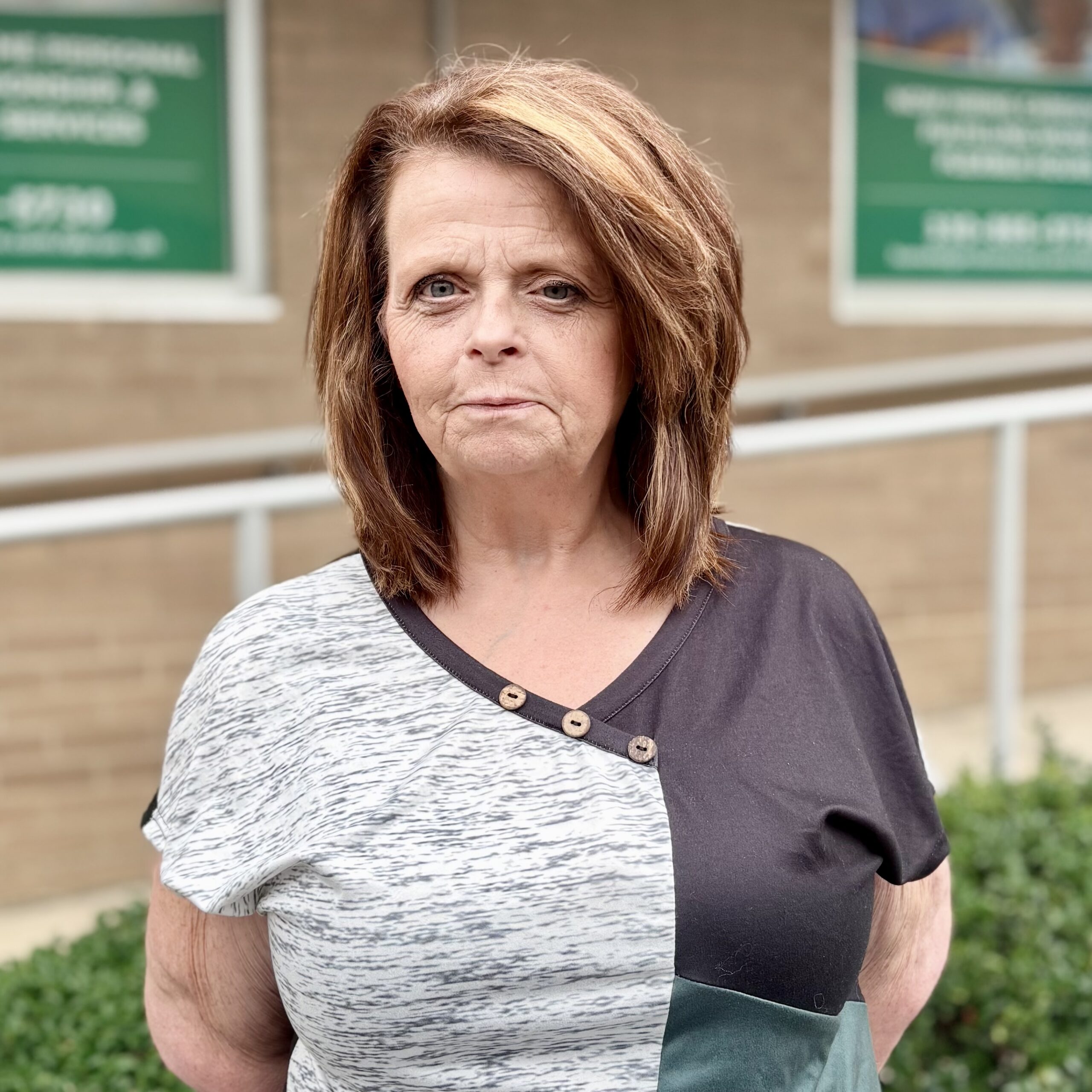 A woman with shoulder-length brown hair stands outside in front of a building with tan bricks and green signs, wearing a color-blocked top with buttons near the collar.