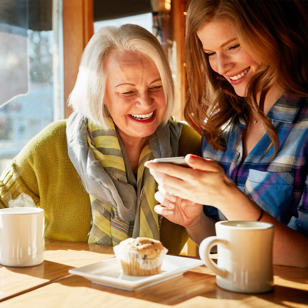 Two women sit at a sunlit table in a café, smiling and looking at a smartphone. There are two mugs and a plate with a muffin on the table, creating a warm and cheerful atmosphere.