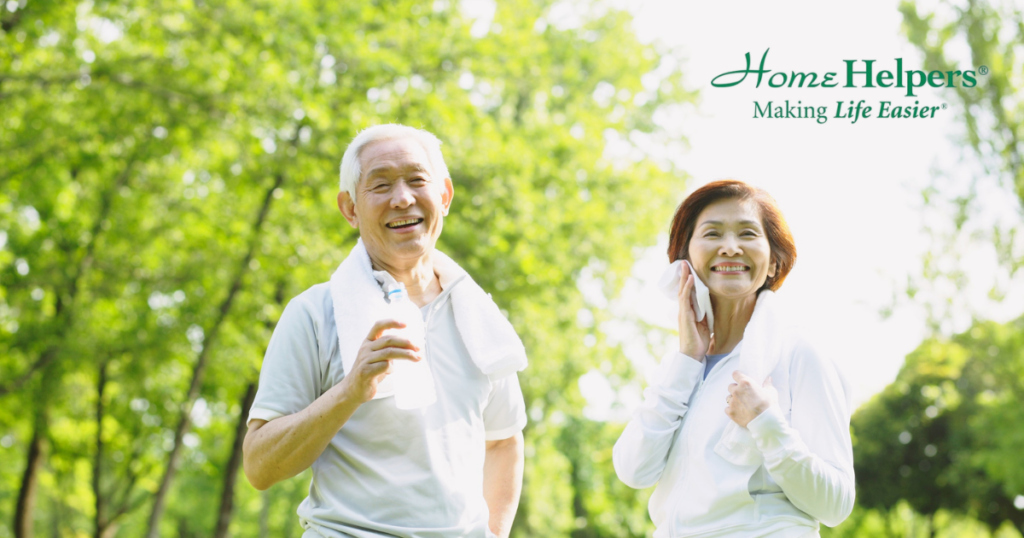Two smiling older adults, a man and a woman, stand outdoors in bright sunlight with towels around their necks. The image includes the Home Helpers logo and the slogan Making Life Easier in the top right corner.