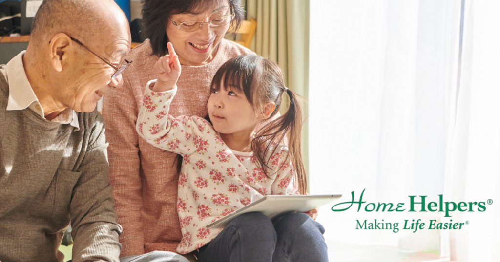 A smiling elderly couple sits with a young girl holding a tablet. The girl gestures upward while they all share a joyful moment. Home Helpers—Making Life Easier text appears in the bottom right.