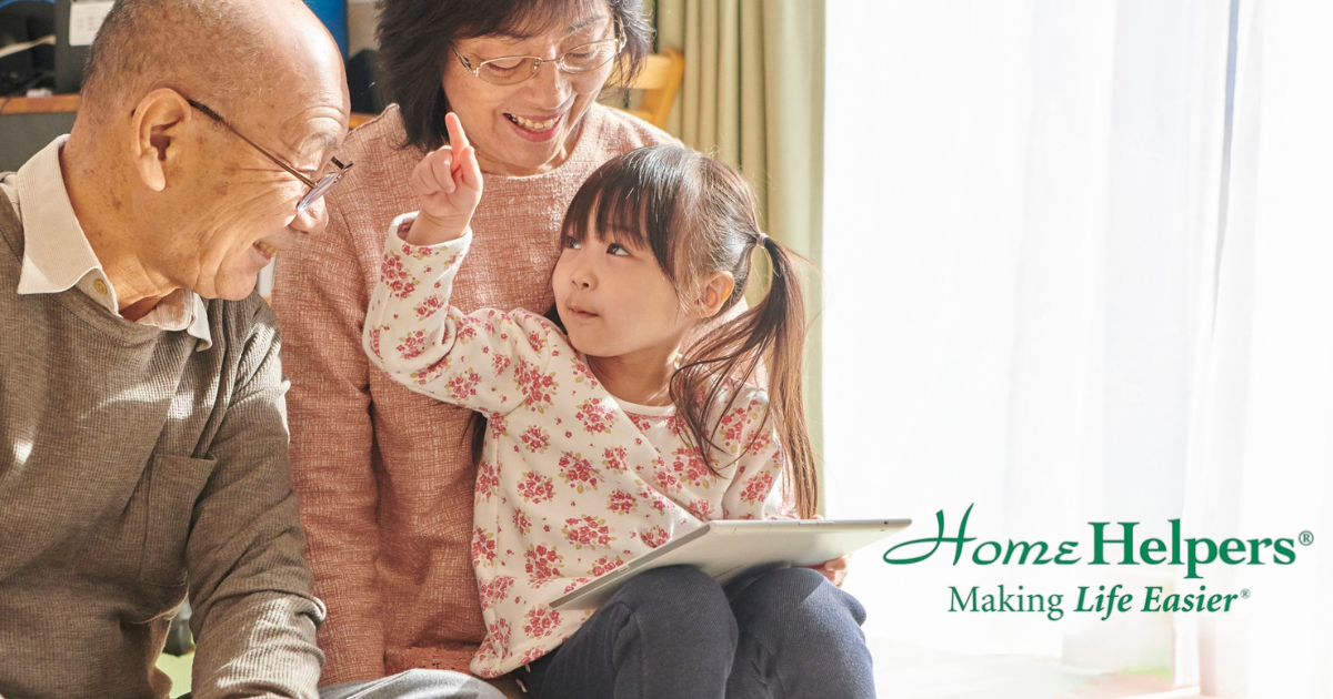 A smiling elderly couple sits with a young girl holding a tablet. The girl gestures upward while they all share a joyful moment. Home Helpers—Making Life Easier text appears in the bottom right.