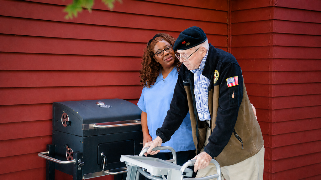 A caregiver in blue scrubs supports an elderly man using a walker as he walks past a grill.