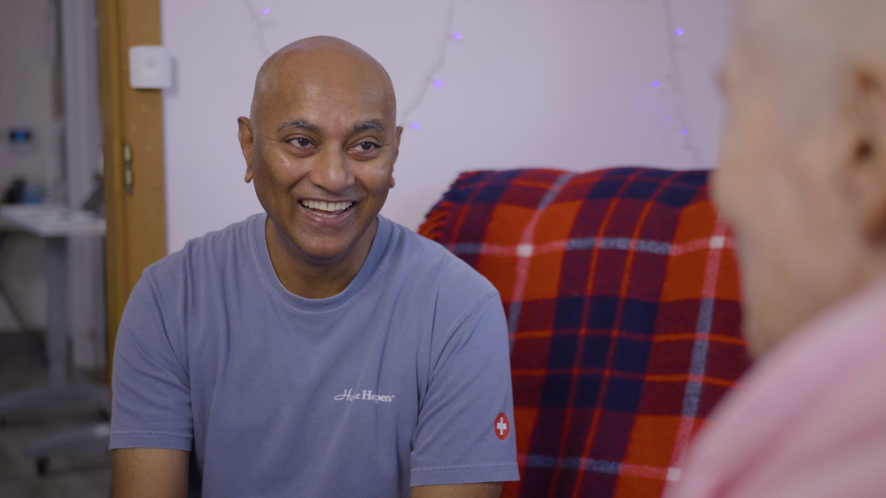A smiling man in a blue t-shirt sits across from another person.