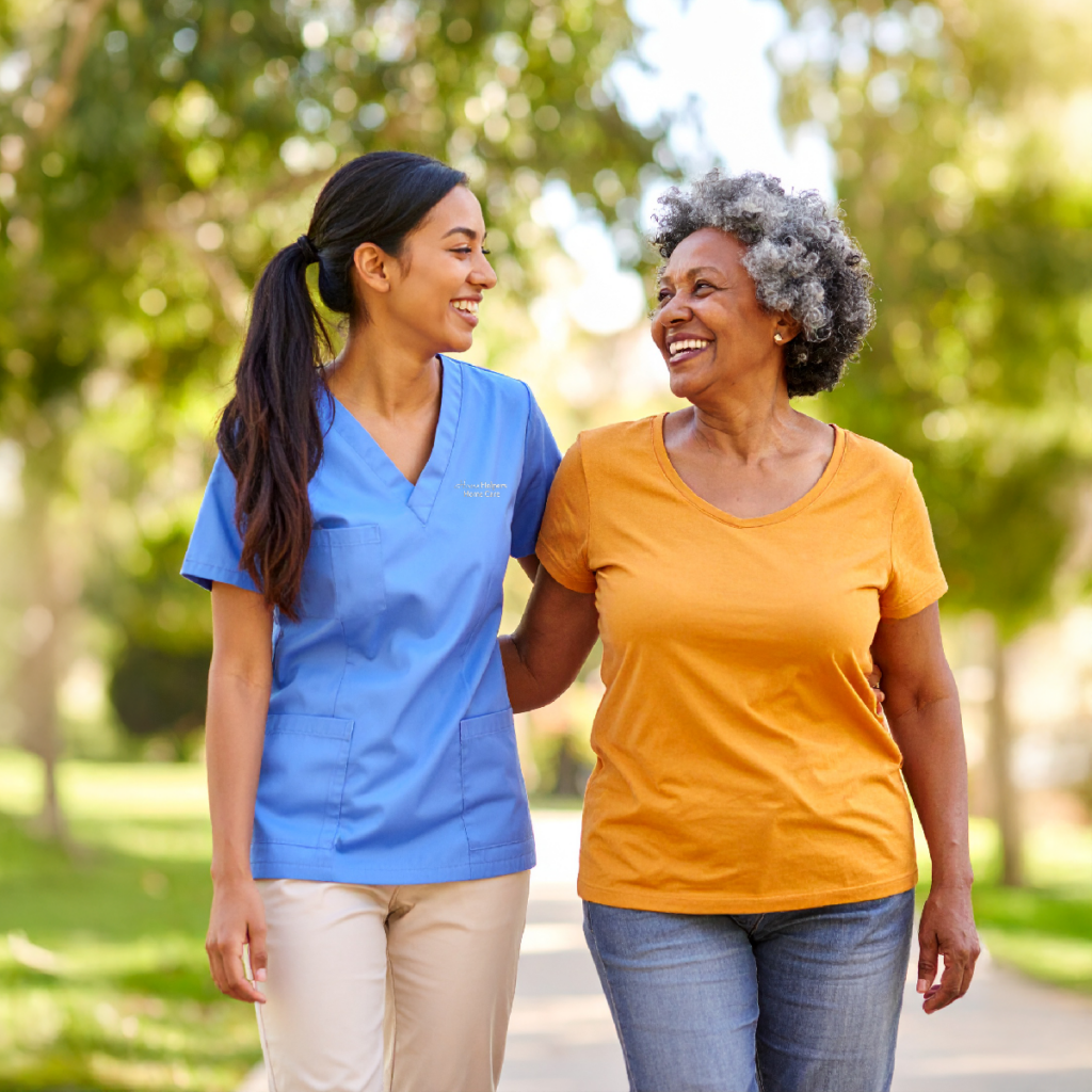 A healthcare worker in blue scrubs walking and laughing with an older woman outdoors.