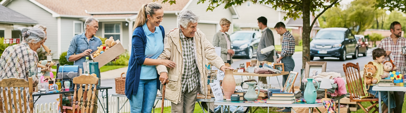 A woman assists an elderly man walking past tables at an outdoor neighborhood yard sale.