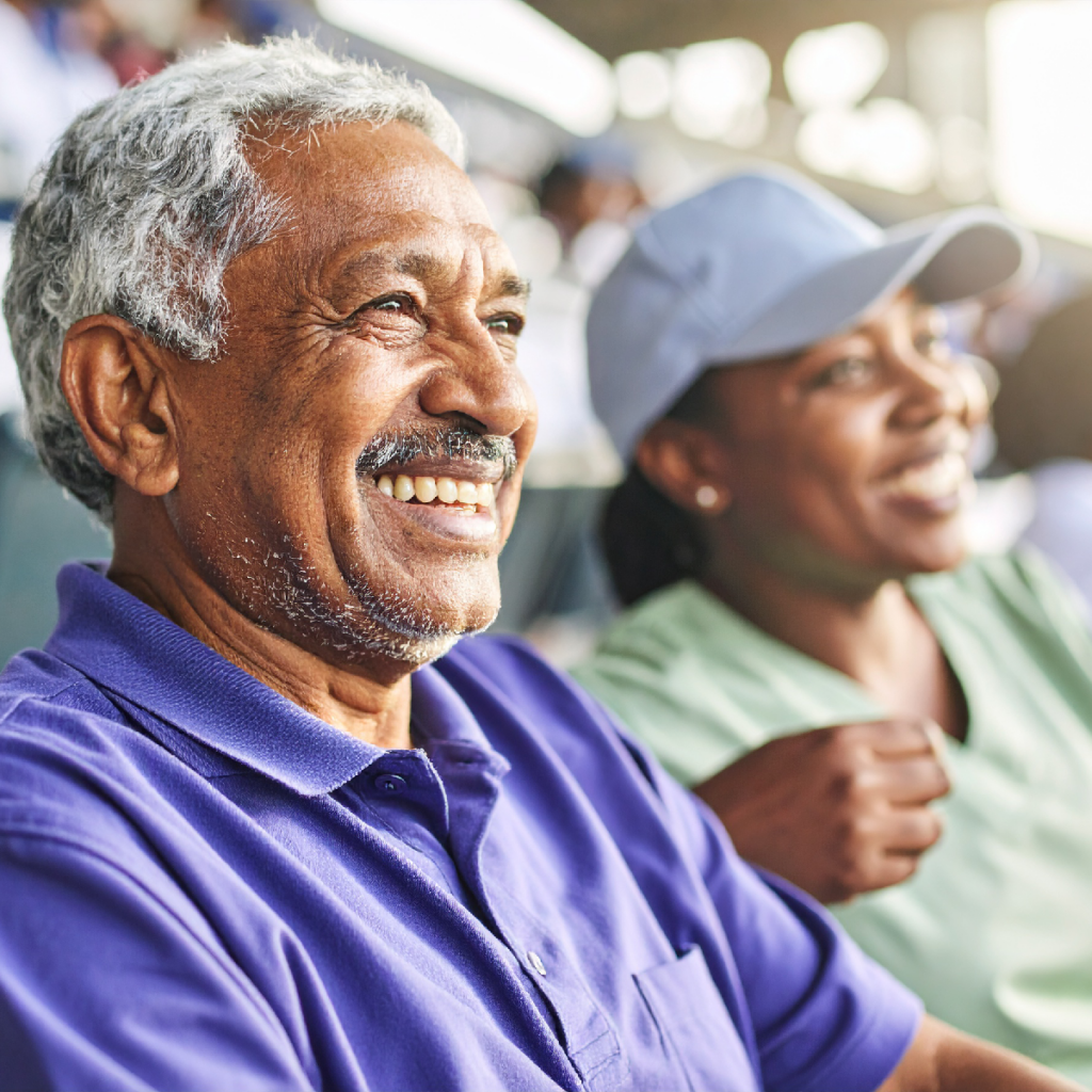 An older man with gray hair and a mustache smiling while sitting next to a woman wearing a baseball cap at a stadium.