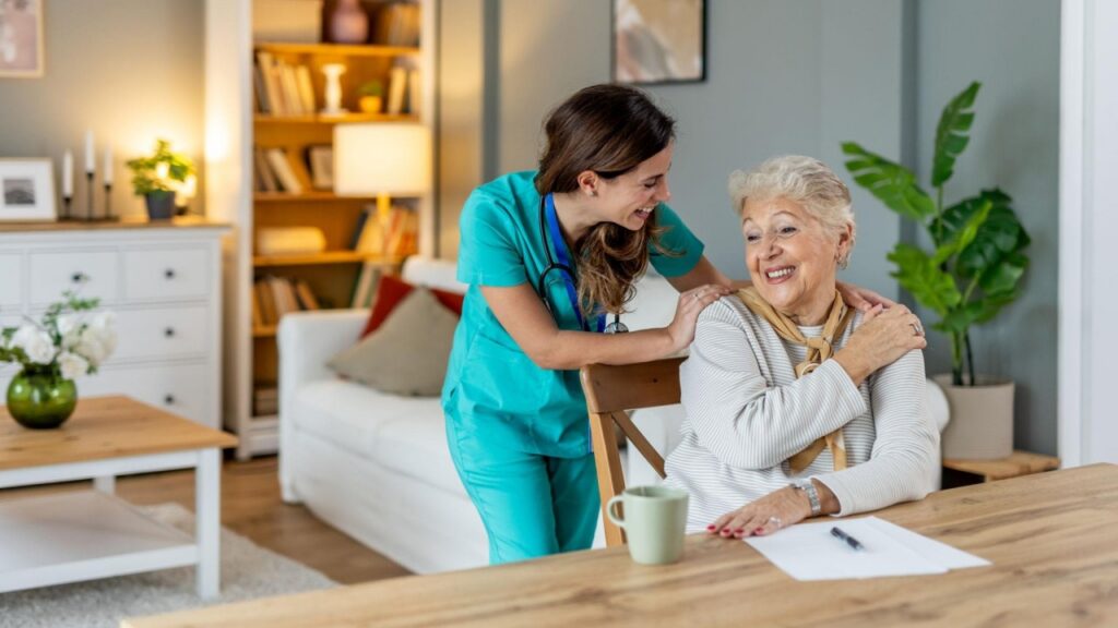 A smiling caregiver in green scrubs rests her hands on the shoulders of an elderly woman seated at a table.