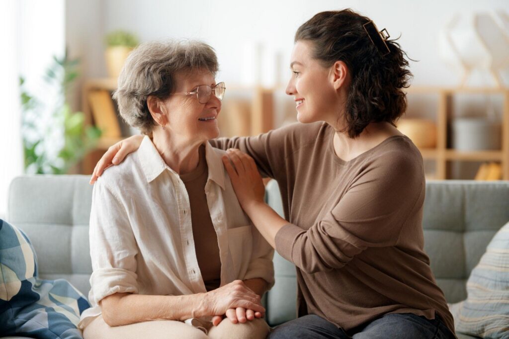 A young woman sits on a sofa with her arm around an elderly woman as they look at each other and smile.