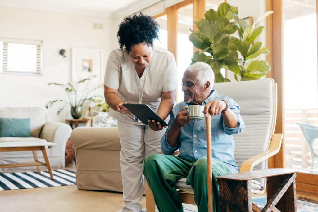 A caregiver shows a tablet to an elderly man sitting in a chair and holding a mug.