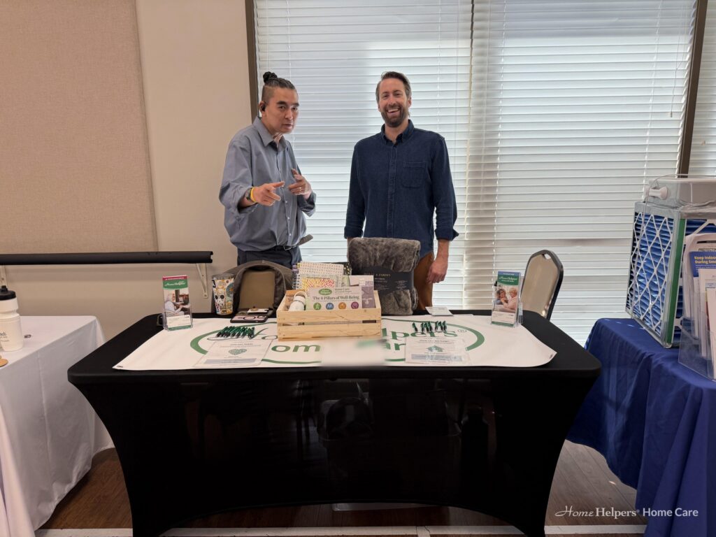 Two men stand behind a trade show table featuring Home Helpers branding and informational pamphlets.