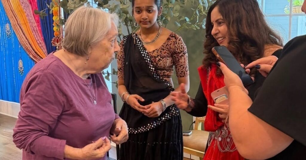 An older woman in a purple shirt converses with a young woman in a black saree and a woman in a red outfit who is holding a smartphone.