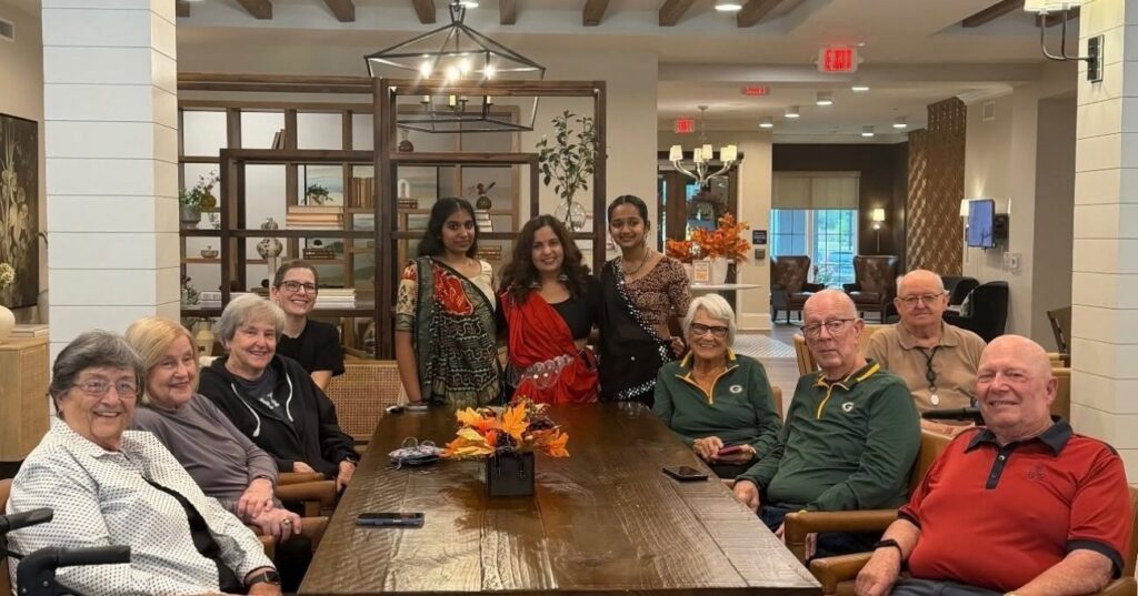 A group of residents and staff members pose for a photo around a long wooden table in a dining area.