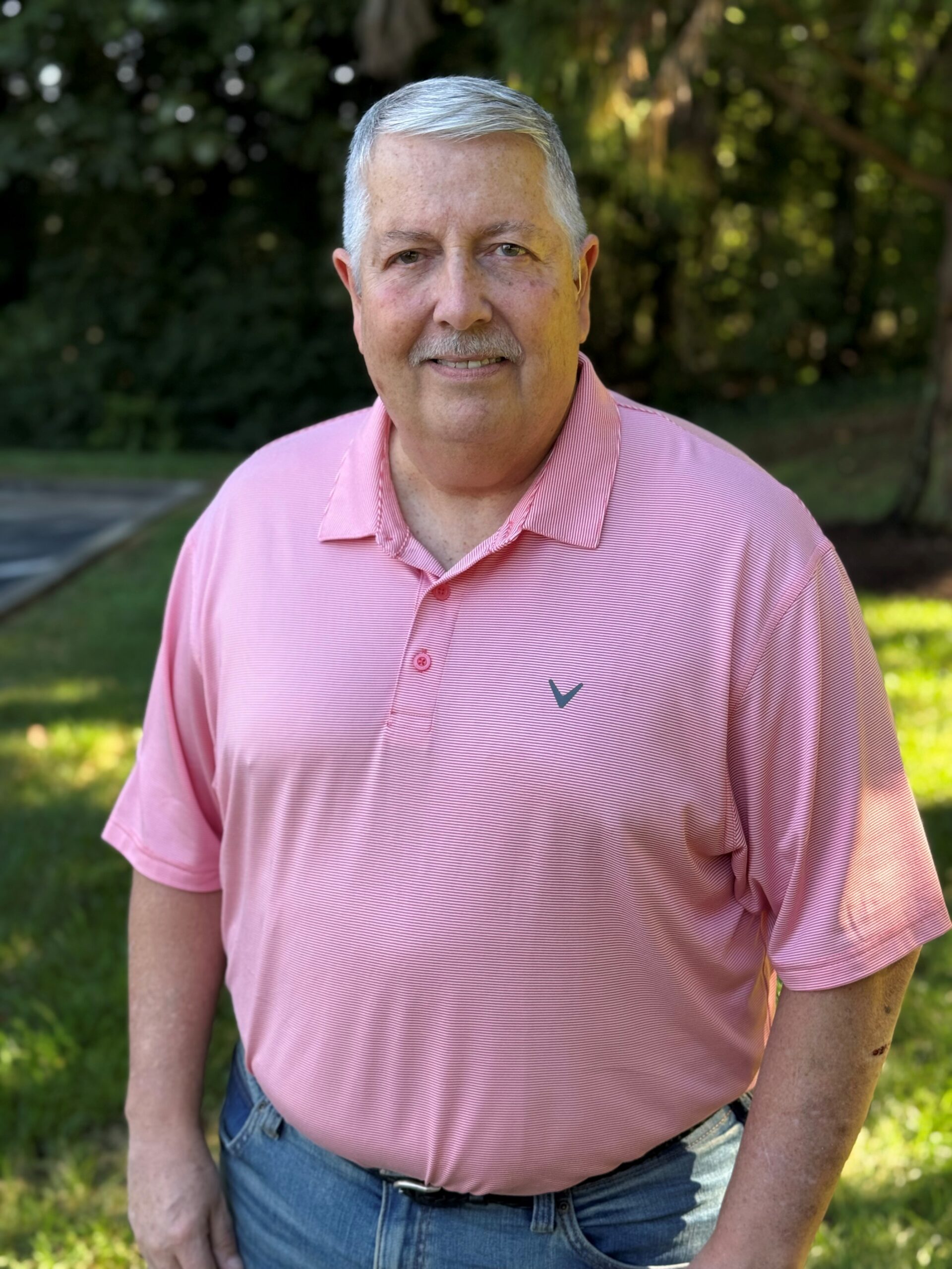 A smiling man with gray hair and a mustache wearing a pink collared shirt.
