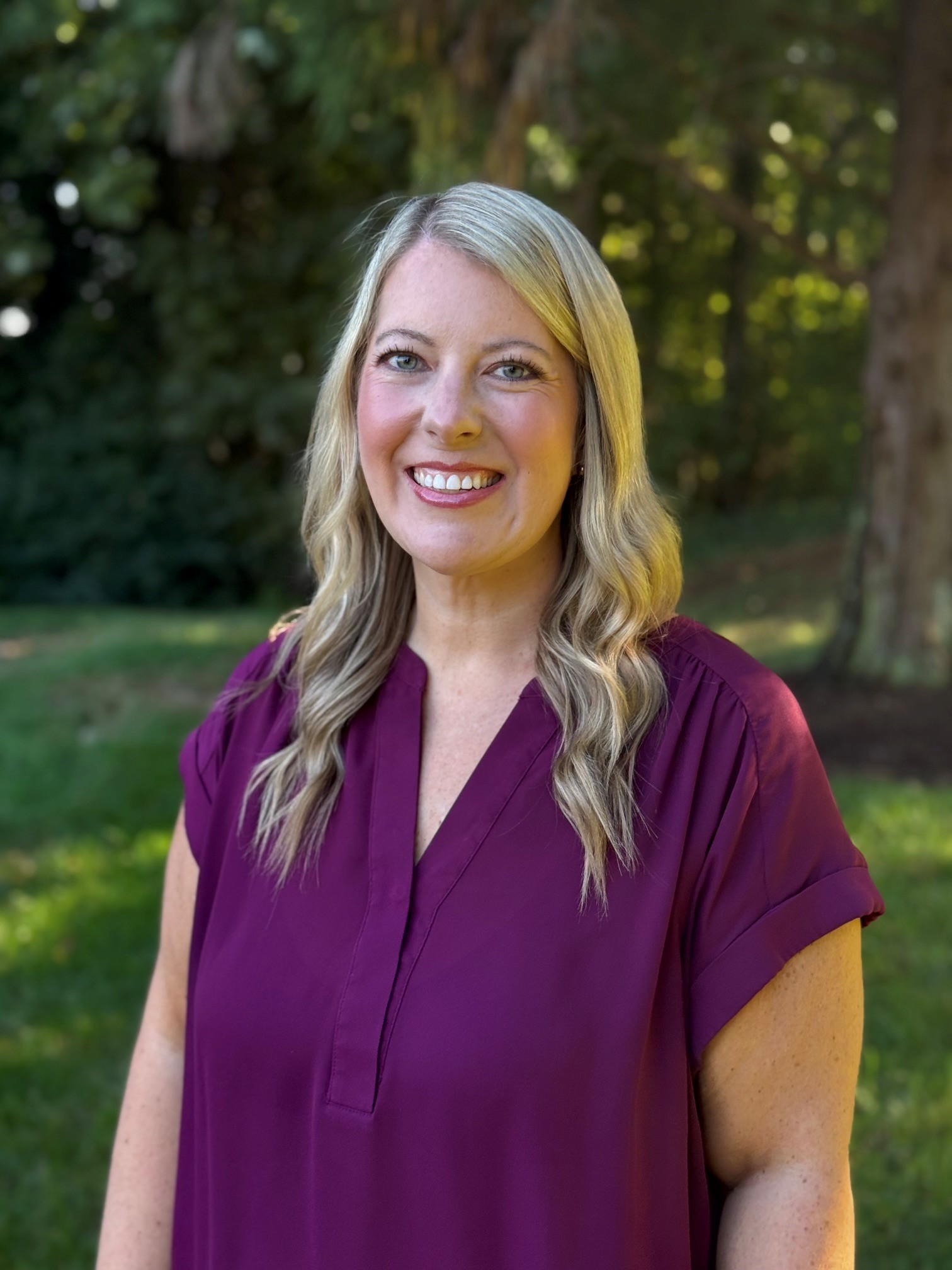 A blonde woman smiling at the camera, wearing a purple shirt outdoors.