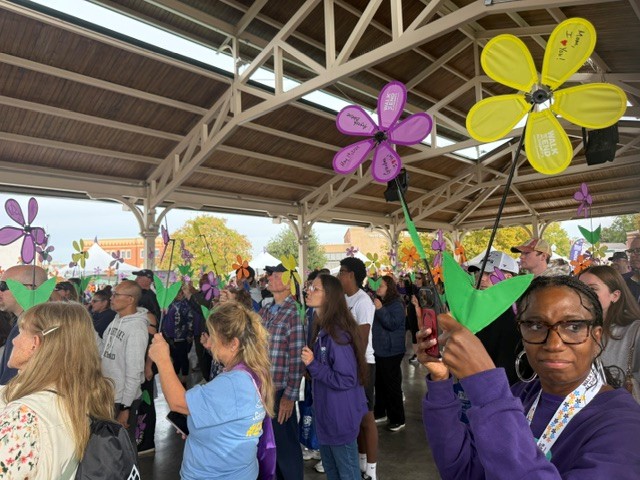 People in an outdoor pavilion holding large, colorful paper flowers on sticks during a community walk event.
