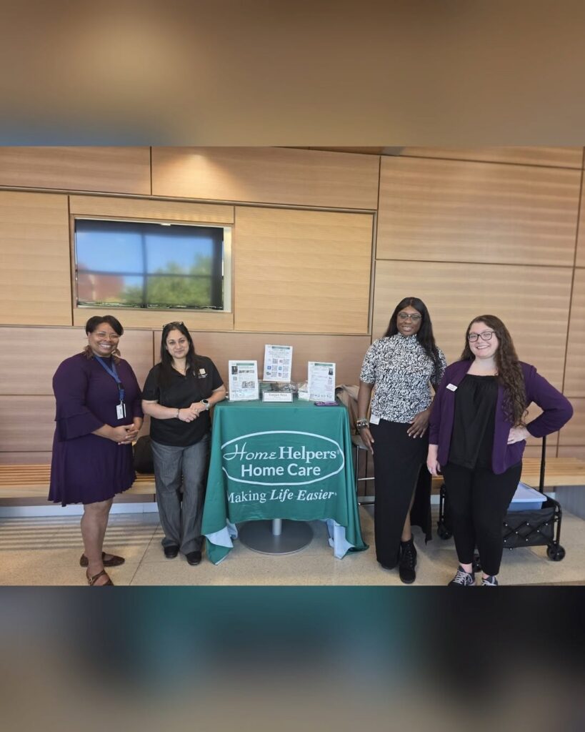 Four people stand behind a green Home Helpers Home Care branded table in an office lobby.