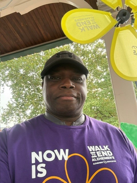 A man wearing a black baseball cap and a purple Walk to End Alzheimers T-shirt stands near a large yellow decorative pinwheel.