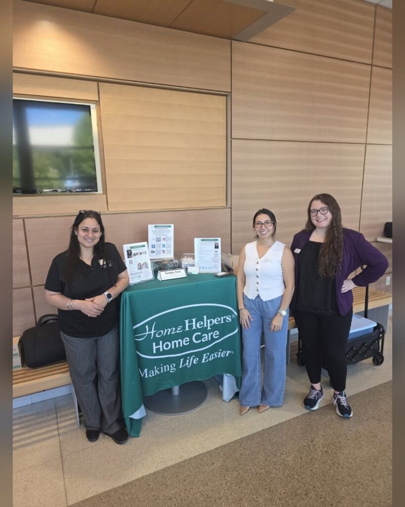 Three women stand behind a Home Helpers Home Care table in an office lobby.