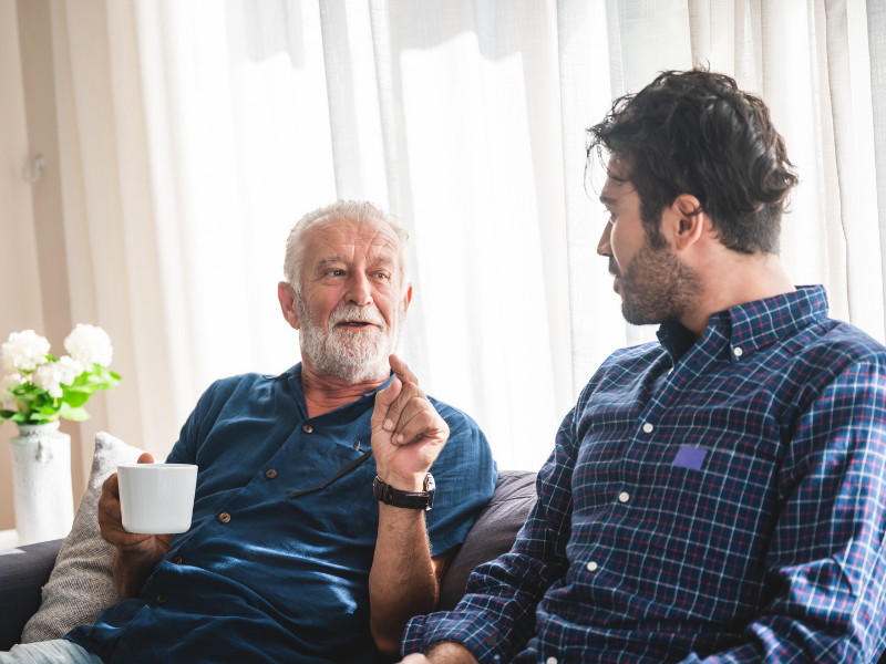 An older man with a white beard holding a mug gestures while talking to a younger man sitting on a sofa.