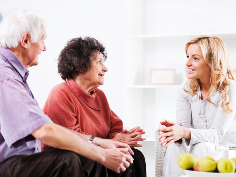An older man and woman sit on a couch, smiling and conversing with a younger woman sitting across from them in a bright, cozy living room.