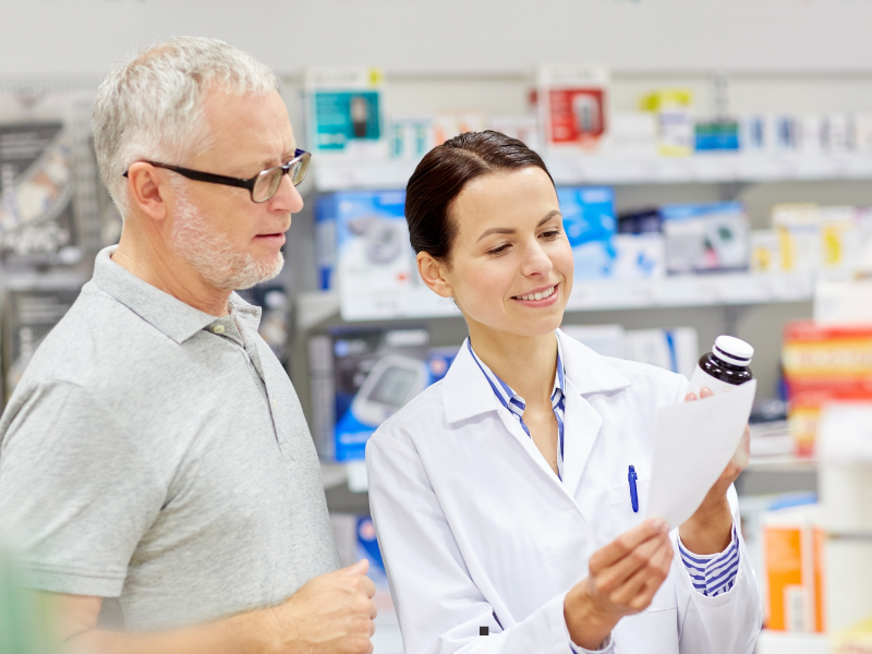 A pharmacist in a white coat shows a bottle of medication and paperwork to an older man in a pharmacy aisle.