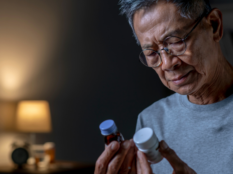 An older man with glasses examines two medication bottles in his hands, appearing focused. A softly lit room with a lamp and a table is blurred in the background.