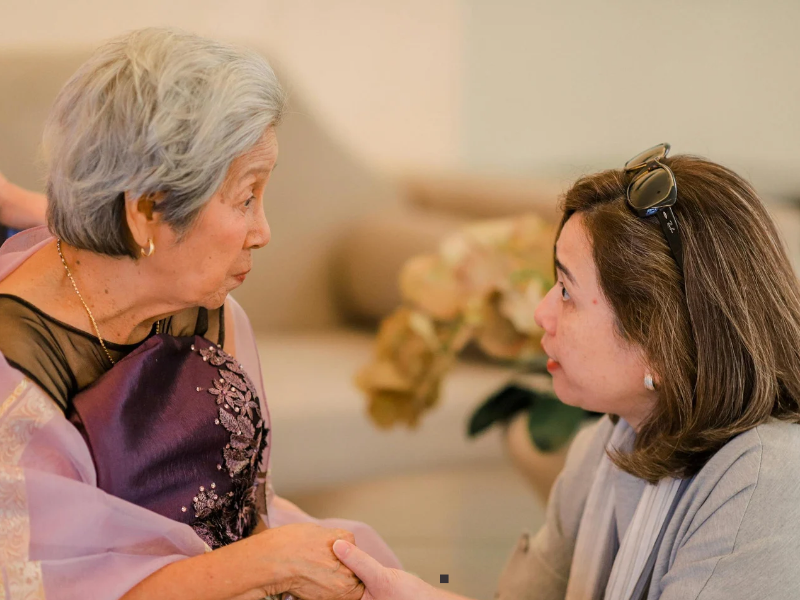 An older woman with gray hair and a younger woman facing her, holding hands and looking at one another.