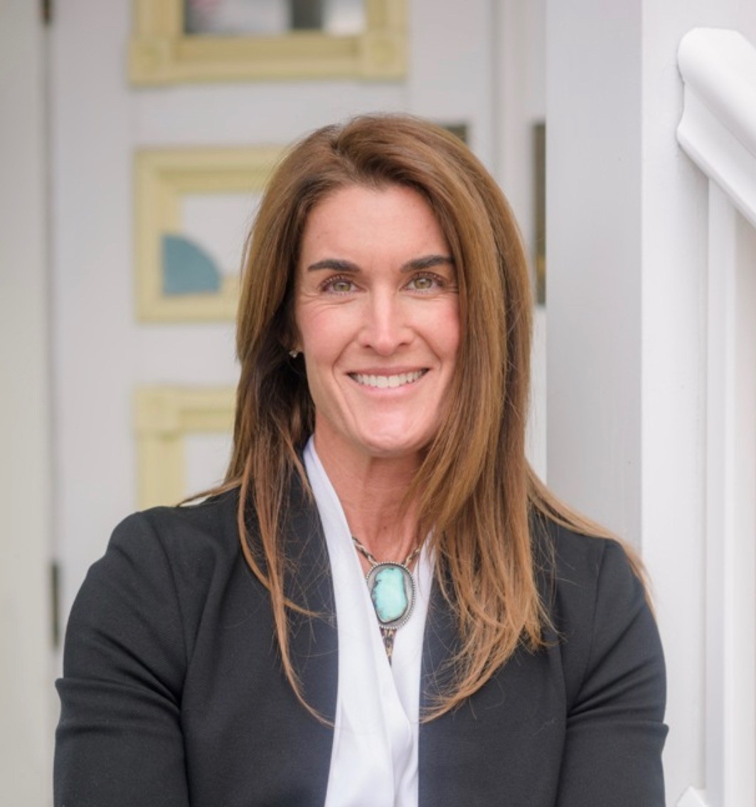 A woman with shoulder-length brown hair smiling, wearing a white shirt, a black blazer, and a turquoise pendant necklace.