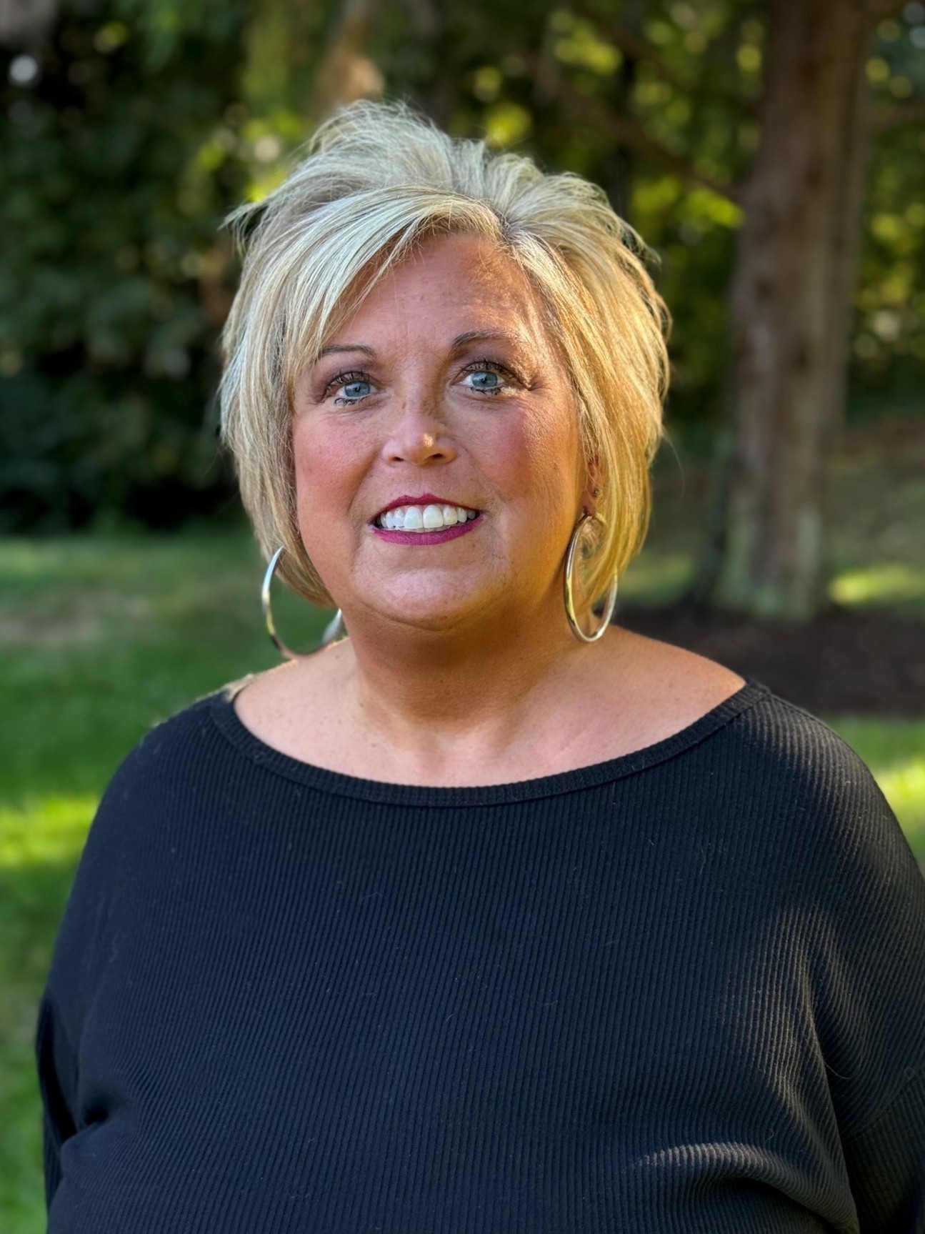 A smiling woman with blonde hair and hoop earrings wearing a black top outdoors.