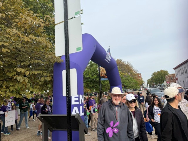 A smiling man wearing a bucket hat stands before a purple Walk to End Alzheimers archway, surrounded by a crowd of participants.