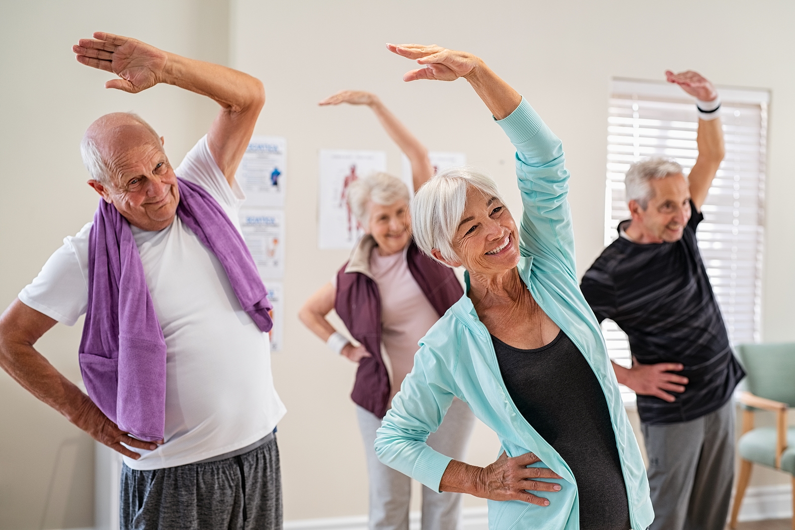 A group of seniors exercising together in a bright room, stretching their arms overhead.