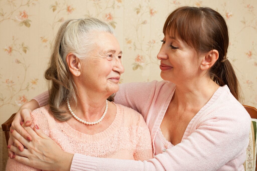 A younger woman smiling and resting her hands on the shoulders of an elderly woman, who is looking back at her.