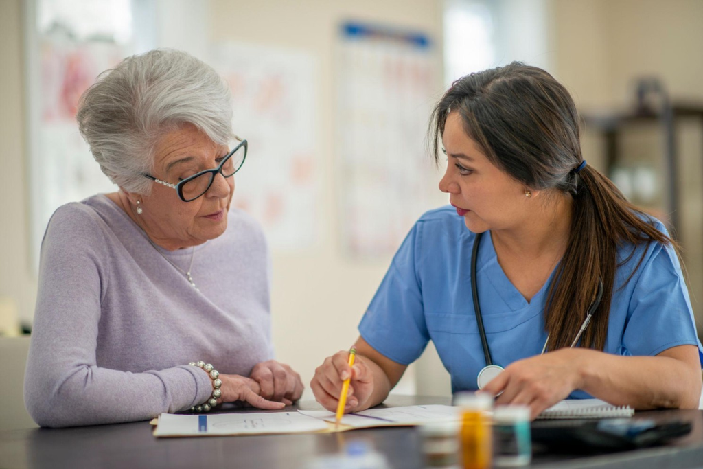 A healthcare worker in blue scrubs sits at a table and reviews medical documents with an elderly woman.