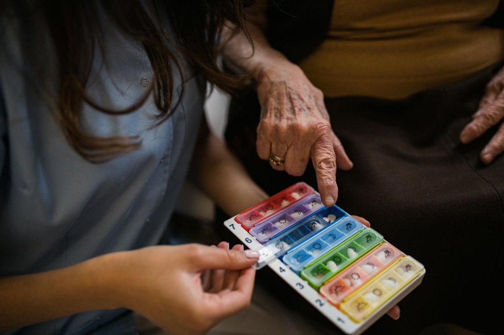 A caregiver helping an older adult open a weekly pill organizer.