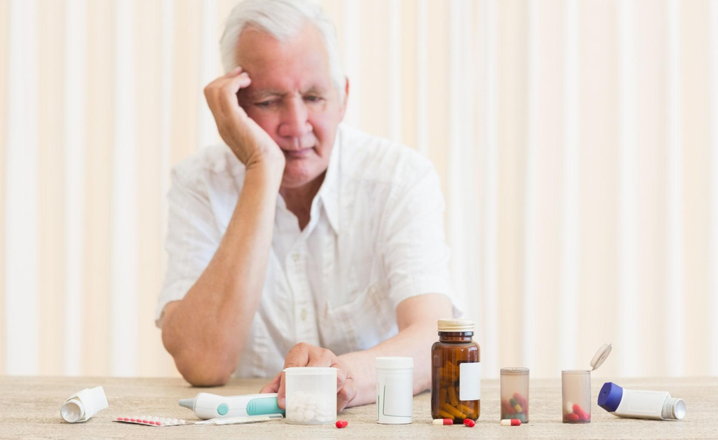 An elderly man sits at a table with his head resting on his hand, looking down at a collection of medication bottles and medical devices.