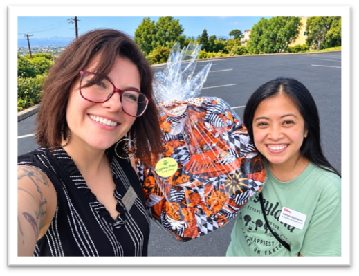 Two women smile while holding a large gift basket wrapped in patterned cellophane.