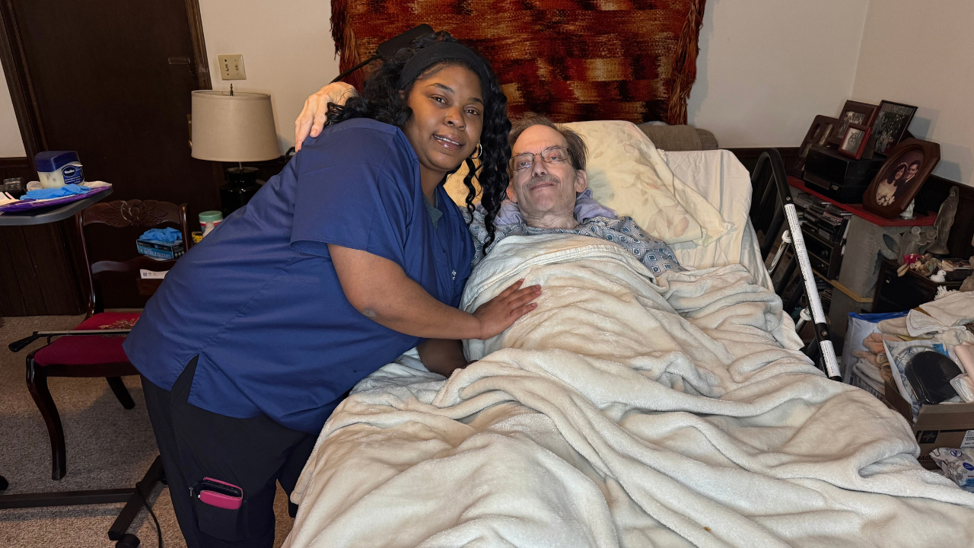A smiling caregiver in blue scrubs with her arm around an elderly man resting in a hospital bed.