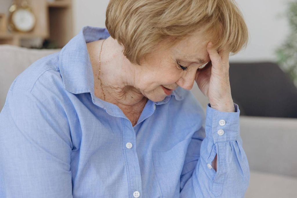 An older woman in a blue collared shirt looking down with her hand on her forehead.