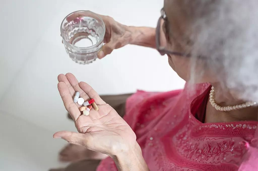 An elderly person holding a glass of water and a handful of assorted pills.